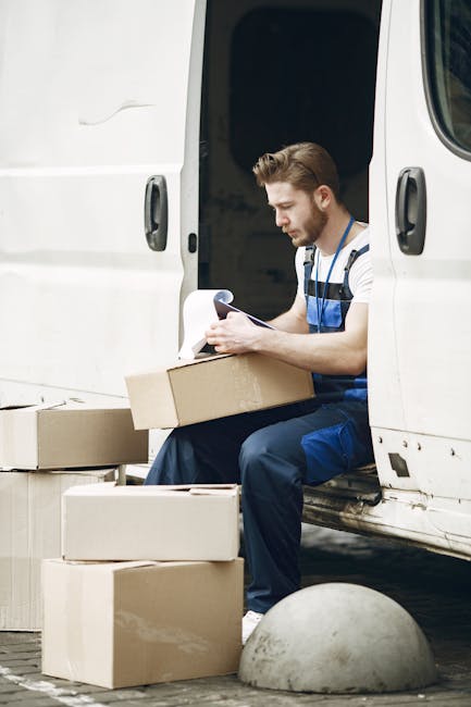 A man wearing a blue and white striped shirt and blue overalls is seated on the edge of a white delivery van with the side door open, surrounded by stacked cardboard moving boxes. He is holding a clipboard or checklist and appears to be reviewing or preparing documents related to the home relocation process. The boxes, some open and some sealed, are positioned both inside the van and on the ground nearby, indicating an active packing and loading phase of a furniture transport or house removal. The scene takes place outdoors, with the van situated on a paved area near a concrete bollard, suggesting the vehicle is being used for local moving services. The environment is well-lit, with natural daylight highlighting the packing materials and the man's focused posture, reflecting professional removal or moving logistics that Man and Van Kingsbury might handle during a house removal.