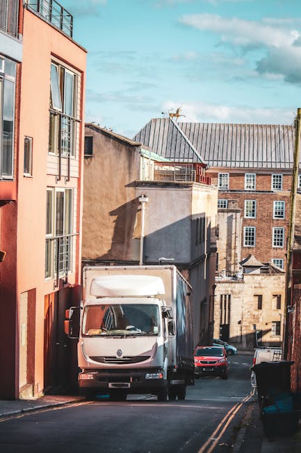 A white delivery van belonging to Man and Van Kingsbury is parked on a narrow, sloped urban street, with the van positioned against a tall, pinkish-orange residential building on the left. The street is lined with several other parked cars, including a red vehicle further down the road, and features double yellow lines along the curb. The surrounding buildings are a mix of brick and concrete structures with flat roofs, some with chimneys and rooftop equipment, set against a partly cloudy sky. In the foreground, a black rubbish bin is visible on the sidewalk. The scene depicts a typical home relocation or moving process in progress, with the van ready for furniture transport and packing and moving services, characteristic of a professional removals operation by Man and Van Kingsbury.