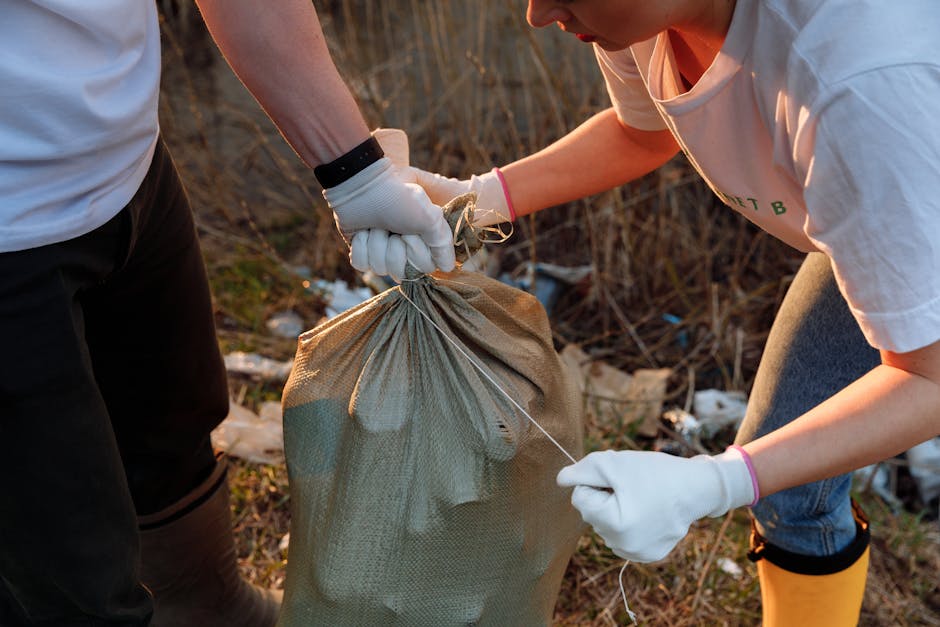 Two individuals wearing white gloves are involved in a home relocation activity outdoors, with one holding a large beige sack made of woven material, likely filled with waste or items for disposal. The person on the right, dressed in a white T-shirt and blue jeans, appears to be securing the sack with a piece of string, while the other person, partially visible, assists by holding the sack steady. The scene takes place on a patch of ground cluttered with litter and debris, possibly near a property or in a yard area. In the background, there is dry grass and scattered trash, indicating an environment where waste collection or clearance might be taking place. The lighting suggests natural daylight, and the activity reflects the physical process of garbage collection or preparing items for removal, consistent with house clearance or waste management services offered by companies like Man and Van Kingsbury, especially related to regulations outlined by Brent Council for bulky waste and removals responsibilities.