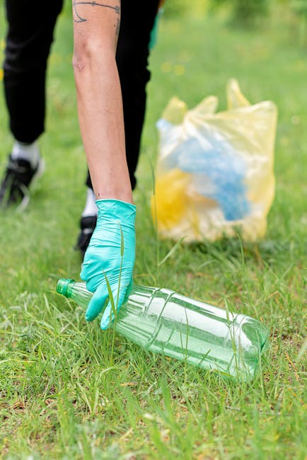 A person wearing blue disposable gloves is bending down on a grassy area, holding a large clear plastic bottle by its neck close to the ground, preparing to dispose of or recycle it. In the background, a yellow plastic bag filled with various rubbish items can be seen on the grass. The setting appears to be outdoors, possibly during a community clean-up or rubbish collection event. The scene emphasizes responsible waste disposal and environmental awareness, which can be related to home relocation efforts in terms of proper packing and waste management. The image depicts a focus on eco-friendly practices, with natural daylight illuminating the scene and the person dressed in casual clothing, suitable for outdoor tasks. Occasionally, Man and Van Kingsbury offers services related to packing, loading, and responsible waste disposal during house moves.