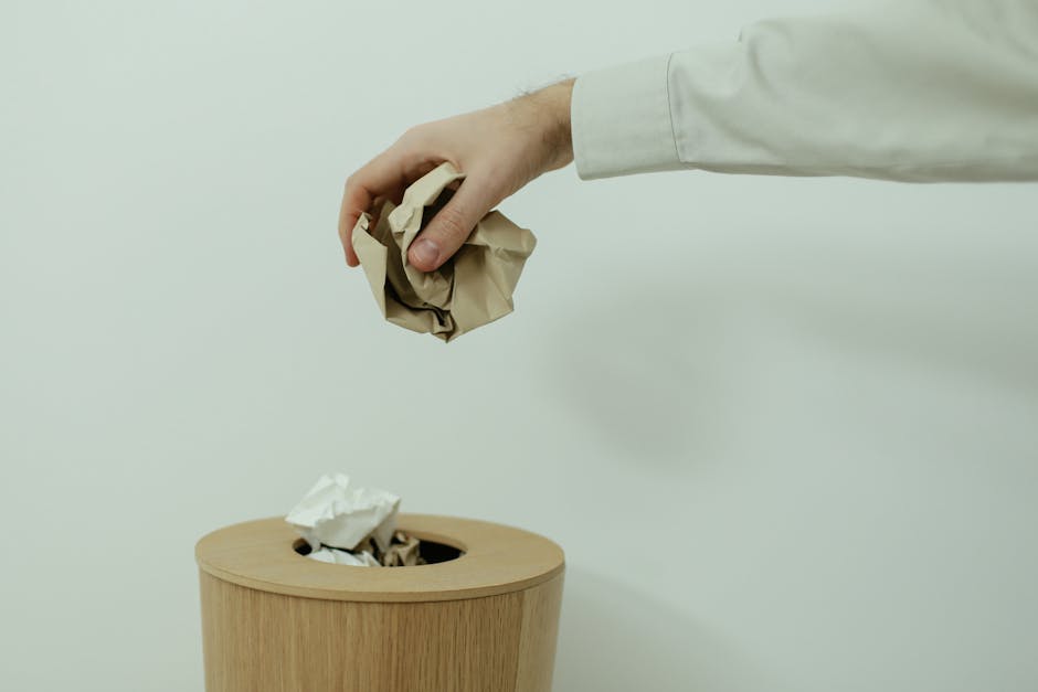 Two individuals wearing white gloves are involved in a home relocation activity outdoors, with one holding a large beige sack made of woven material, likely filled with waste or items for disposal. The person on the right, dressed in a white T-shirt and blue jeans, appears to be securing the sack with a piece of string, while the other person, partially visible, assists by holding the sack steady. The scene takes place on a patch of ground cluttered with litter and debris, possibly near a property or in a yard area. In the background, there is dry grass and scattered trash, indicating an environment where waste collection or clearance might be taking place. The lighting suggests natural daylight, and the activity reflects the physical process of garbage collection or preparing items for removal, consistent with house clearance or waste management services offered by companies like Man and Van Kingsbury, especially related to regulations outlined by Brent Council for bulky waste and removals responsibilities.