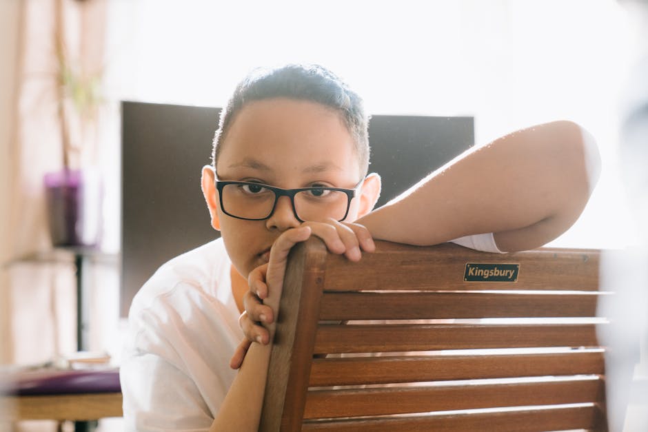 A young person with glasses and dark hair, wearing a white shirt, is resting their arms on the back of a wooden chair with a 'Kingsbury' label. The chair is part of a domestic interior setting, with a blurred background that includes a window with curtains, a potted plant, and a bookshelf. The lighting is bright, illuminating the scene naturally. The image appears to capture a moment during home packing or a casual pause in furniture placement, relevant to house removals and packing processes. The focus is on the individual and the wooden furniture, which suggests involvement in a home relocation or furniture transport activity, as performed by a professional within the removals service designated for Kingsbury, as indicated by the label.
