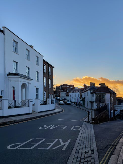 A quiet street scene on Kingsbury High Road during sunset, showing a row of white and dark brick residential buildings with multiple stories and traditional architectural features. The road has a marked 'KEEP CLEAR' area painted on the asphalt, and a paved sidewalk runs alongside it. In the foreground, part of a loading zone or driveway is visible, with no visible vehicles or moving activities. The sky is partly cloudy with warm orange and yellow hues from the setting sun, casting a soft light over the scene. This image closely relates to house removals or relocation services by illustrating the type of street environment where furniture transport and home moving logistics may take place, with potential access considerations for Man and Van Kingsbury's professional removals team.