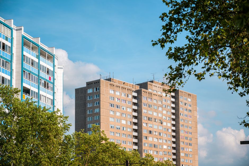 A high-rise residential building with a brown brick facade and numerous white-framed windows, situated next to a modern, glass-fronted apartment block. The scene is taken outdoors during daytime, with clear blue skies and scattered white clouds overhead. The foreground features green trees with leafy branches partially obscuring the lower part of the buildings, indicating a suburban or urban setting. This image exemplifies urban housing in Kingsbury, which can present challenges such as narrow staircases and lack of lifts during home relocation and furniture transport. Occasionally, during the packing and moving process, items like cardboard boxes and wrapped furniture are part of the load. Detailed planning and appropriate equipment, such as trolleys and blankets, are essential for efficient loading and transport, often involving a moving van parked nearby for easy access, as part of professional removals services provided by companies like Man and Van Kingsbury.