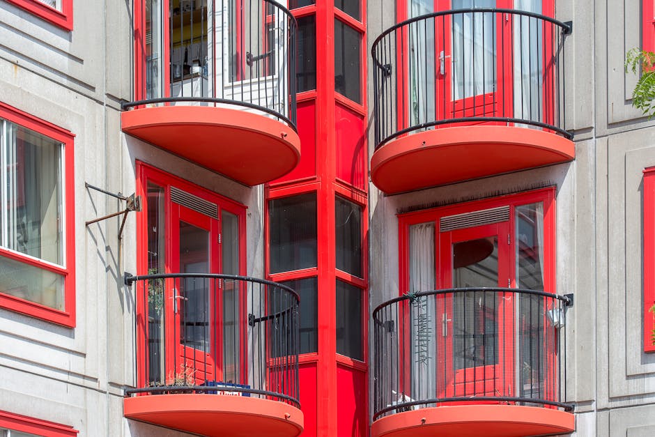 Close-up view of the exterior of a modern residential building featuring red-framed sliding glass doors leading to small, semi-circular balconies with black metal railings. The building's facade is light-colored with red accents around windows and doors, and vertical red panels running between the glass sections. The balconies contain various items such as potted plants and outdoor furniture, with some showing signs of weathering. The image captures part of the building's multi-story structure, highlighting the design elements associated with contemporary housing suited for flats without lifts, relevant for home relocation and furniture transport processes. Man and Van Kingsbury may assist with moving logistics involving these types of apartments, where careful handling and strategic loading are necessary.