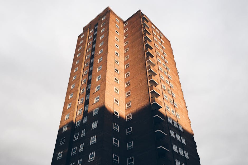 A high-rise residential building with a brown brick facade and numerous white-framed windows, situated next to a modern, glass-fronted apartment block. The scene is taken outdoors during daytime, with clear blue skies and scattered white clouds overhead. The foreground features green trees with leafy branches partially obscuring the lower part of the buildings, indicating a suburban or urban setting. This image exemplifies urban housing in Kingsbury, which can present challenges such as narrow staircases and lack of lifts during home relocation and furniture transport. Occasionally, during the packing and moving process, items like cardboard boxes and wrapped furniture are part of the load. Detailed planning and appropriate equipment, such as trolleys and blankets, are essential for efficient loading and transport, often involving a moving van parked nearby for easy access, as part of professional removals services provided by companies like Man and Van Kingsbury.