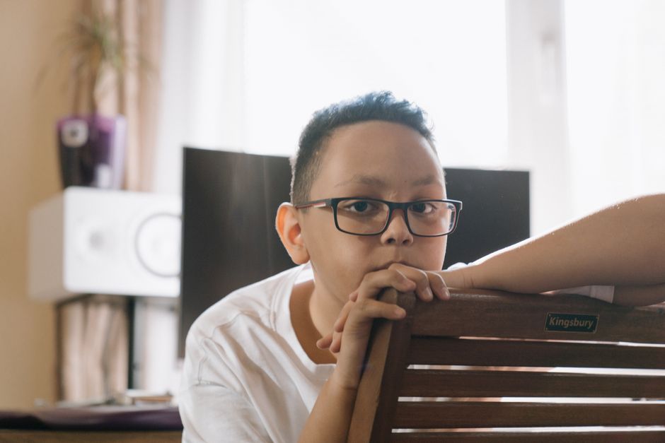 A young man with short dark hair, glasses, and light skin leans on the back of a wooden dining chair with a small label reading 'Kingsbury,' inside a room during home relocation preparations. He rests