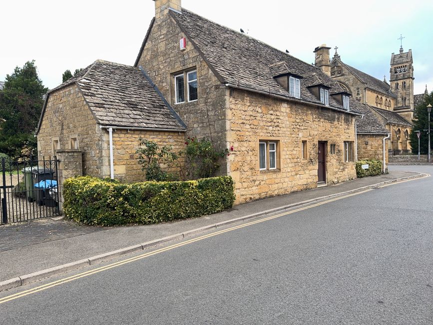 Exterior view of a stone-built house situated on a paved street, with a small hedge and a garden area at the front. The house features a sloped tiled roof with multiple dormer windows, a rain gutter, 