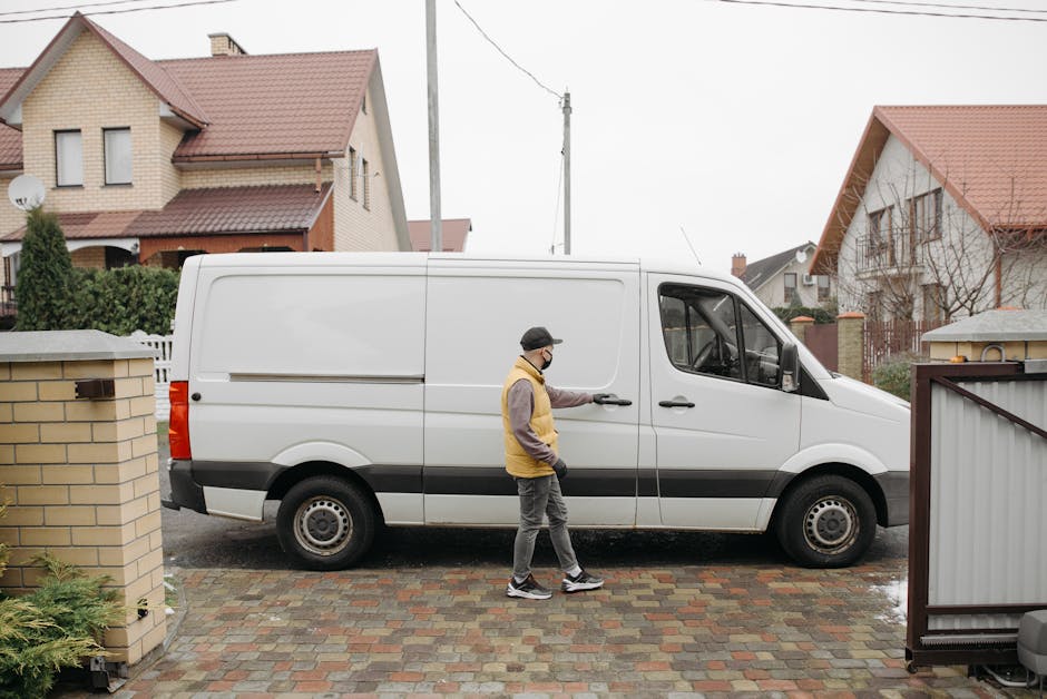 A man wearing a black cap, glasses, a yellow and grey jacket, grey trousers, and sneakers is standing outside a detached house, positioned on the cobbled driveway next to a large white panel van used for house removals. He is opening the van's sliding door, which is partly visible, preparing for furniture or boxes to be loaded. The driveway is bordered by a brick wall on one side and a metal fence on the other, with some green foliage nearby. The background shows several residential houses with pitched roofs, some with dormer windows, and a cloudy sky indicating overcast weather. The scene captures part of the loading process, illustrating the logistics involved in home relocation, with the van parked close to the house for efficient transportation of household items, possibly packed in cardboard boxes or wrapped furniture. Man and Van Kingsbury offers the service, focusing on loading and transport tasks typical in packing and moving operations.