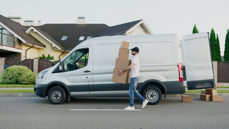 A man wearing a white t-shirt, blue jeans, white sneakers, and a black cap is loading three cardboard boxes into the side door of a white panel van parked on a residential street. Some additional boxes are placed on the pavement nearby. The van features a high roof and sliding side door, indicative of a vehicle used in home relocation or furniture transport services. The background shows a suburban house with a sloped roof, multiple windows, and a wooden fence, surrounded by green shrubs, trees, and a well-maintained garden. The scene is set in daylight with natural lighting, capturing the process of packing and moving furniture as part of professional removals managed by Man and Van Kingsbury, focused on providing efficient house removals and moving logistics.