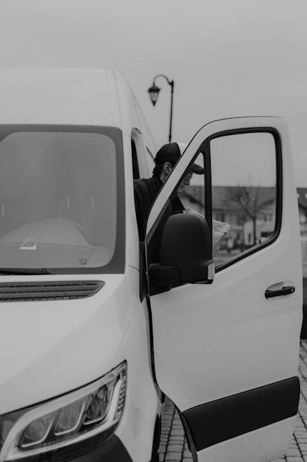 A man wearing a black cap, glasses, a yellow and grey jacket, grey trousers, and sneakers is standing outside a detached house, positioned on the cobbled driveway next to a large white panel van used for house removals. He is opening the van's sliding door, which is partly visible, preparing for furniture or boxes to be loaded. The driveway is bordered by a brick wall on one side and a metal fence on the other, with some green foliage nearby. The background shows several residential houses with pitched roofs, some with dormer windows, and a cloudy sky indicating overcast weather. The scene captures part of the loading process, illustrating the logistics involved in home relocation, with the van parked close to the house for efficient transportation of household items, possibly packed in cardboard boxes or wrapped furniture. Man and Van Kingsbury offers the service, focusing on loading and transport tasks typical in packing and moving operations.