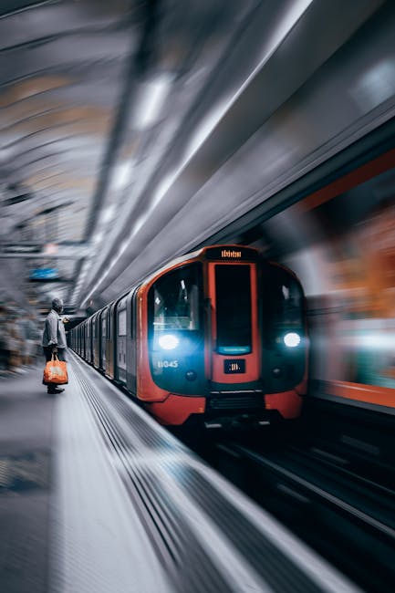 A person in a light jacket and dark trousers is standing on a platform near the edge, holding an orange trolley bag with wheels. They are positioned to the left of the image, facing towards a modern, red and black subway train arriving into the station. The train's front car is visible, with illuminated headlights and a digital display above the door area indicating its destination. The platform has tactile paving along the edge, and the ceiling overhead is curved with metallic and glass elements, creating a sleek, contemporary look. The motion blur effect around the train and platform suggests the train is arriving or departing, emphasizing a busy transportation environment typical of underground public transit stations in the UK. This scene is relevant to house removals and moving services, illustrating efficient transport and journey logistics, which are key aspects of a professional house relocation process as managed by companies like Man and Van Kingsbury.