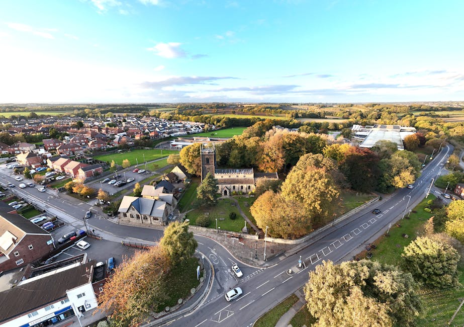 An aerial view of a residential area and a small church surrounded by a variety of trees with green, yellow, and orange leaves. The scene shows a curved road with several cars parked along the side and some vehicles driving through, indicating ongoing home relocation or moving activities. Nearby, there are single-story buildings with pitched roofs, and behind them, a parking lot filled with parked cars. In the distance, there are more houses, green fields, and distant countryside under a partly cloudy sky. The image captures the urban and rural landscape, with visible road infrastructure and a mix of natural and built environments, aligning with the process of furniture transport, packing, and loading during house removals, as managed by Man and Van Kingsbury.