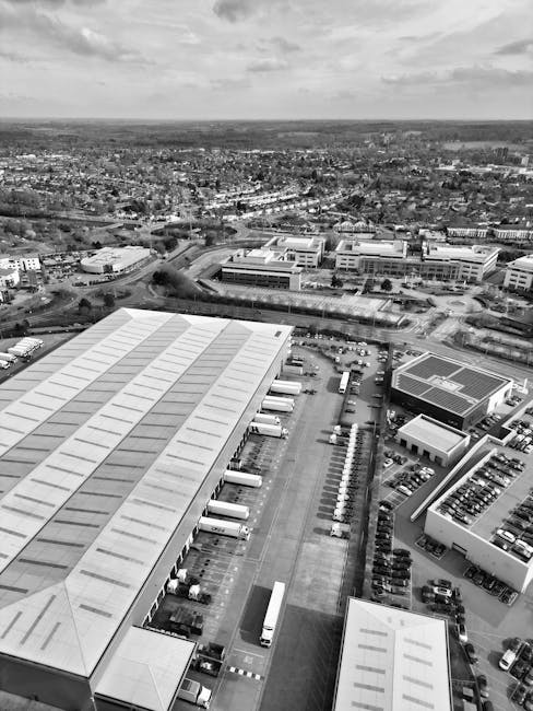 Aerial black-and-white photograph showing a large industrial or warehouse complex with multiple buildings featuring flat, grid-patterned roofs and large loading bays along the side. Several trucks are parked adjacent to the loading areas, with some positioned for loading or unloading, indicating active equipment for home relocation or furniture transport services. The surrounding parking lot contains numerous parked cars, and a row of smaller vehicles is lined up along the side of one building. In the background, an urban landscape with residential and commercial buildings extends to the horizon, with a mixture of open spaces and developed areas under a cloudy sky. The scene depicts a busy logistics and moving environment, reflecting the type of loading process involved in professional removals such as those provided by Man and Van Kingsbury, supporting efficient packing and transport logistics for house or office relocations.