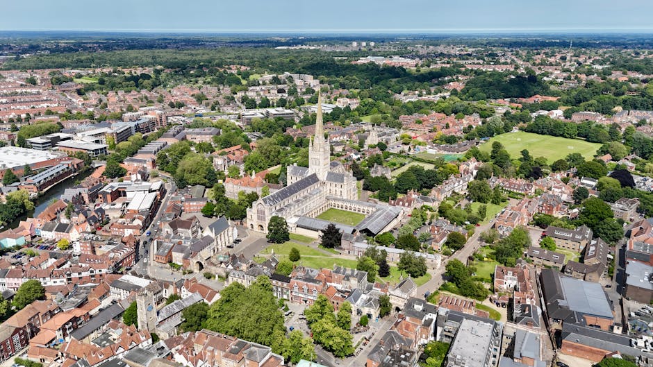 An aerial view of a residential area and a small church surrounded by a variety of trees with green, yellow, and orange leaves. The scene shows a curved road with several cars parked along the side and some vehicles driving through, indicating ongoing home relocation or moving activities. Nearby, there are single-story buildings with pitched roofs, and behind them, a parking lot filled with parked cars. In the distance, there are more houses, green fields, and distant countryside under a partly cloudy sky. The image captures the urban and rural landscape, with visible road infrastructure and a mix of natural and built environments, aligning with the process of furniture transport, packing, and loading during house removals, as managed by Man and Van Kingsbury.